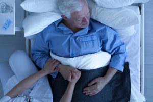 Doctor or nurse holding hand of senior male patient lying in hospice bed